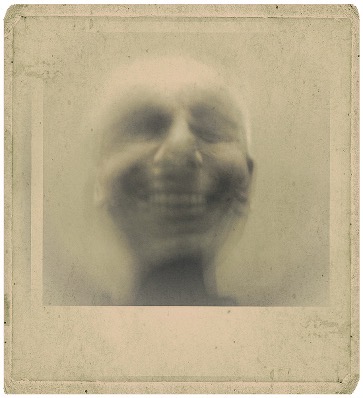 Blurry greyscale image of a person smiling widely, creating an eerie and ghostly effect. The vintage photo border adds an aged, mysterious tone.